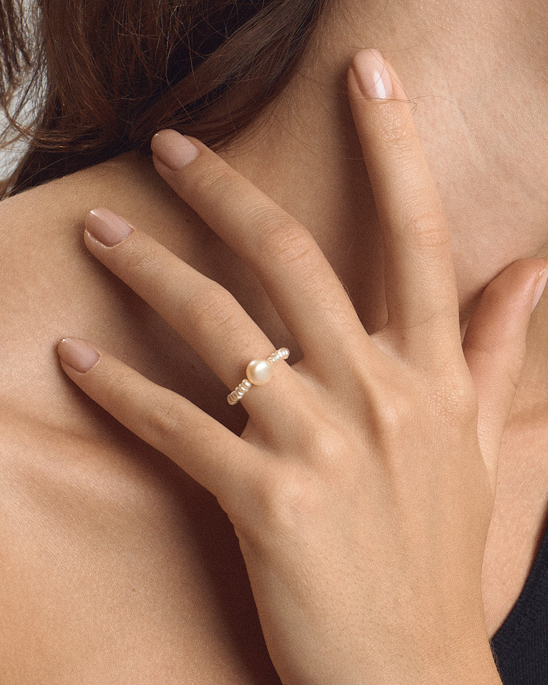 Female model wearing PEARL NAKED Bubble Ring in Pearls, gold-toned beaded band with a central pearl on her finger, hand resting against her neck, for COLD SKIN.